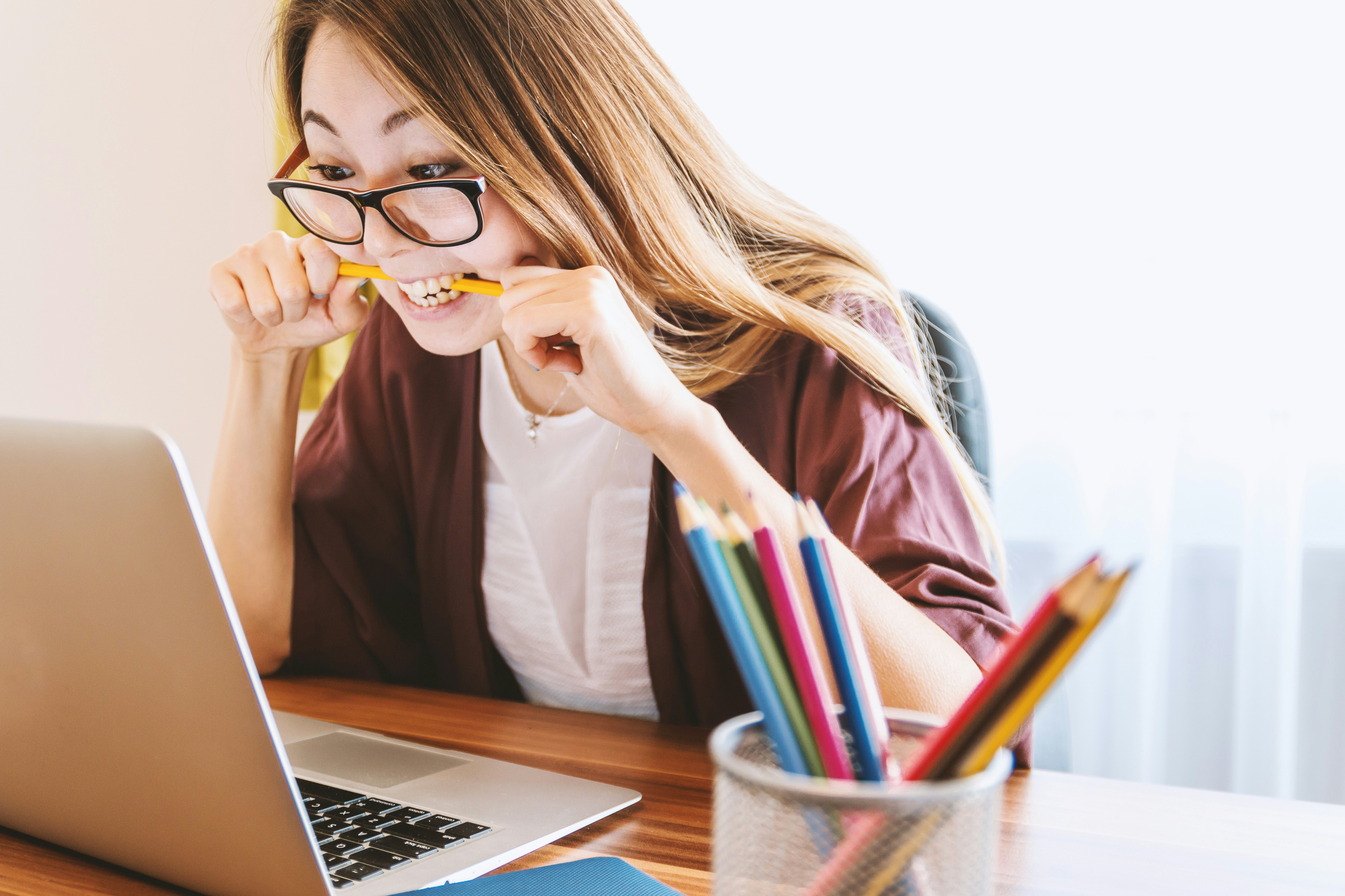 A woman wearing glasses bites a pencil while looking at a laptop screen. She is seated at a desk with a container of colored pencils and a notebook, possibly planning how to leverage email to share news for her small business.