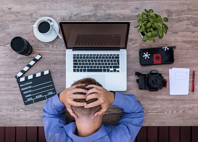 A person sits with their hands on their head in front of a laptop on a wooden desk, surrounded by filmmaking equipment, a coffee cup, a plant, and a notebook. They appear deep in thought as if uncovering the secrets to email success amidst this creative chaos.