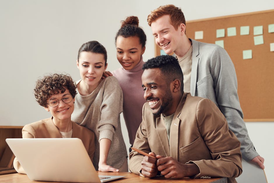 Five people are gathered around a laptop, looking at the screen and smiling, suggesting a collaborative work environment or a group discussion. A corkboard with sticky notes is visible in the background.