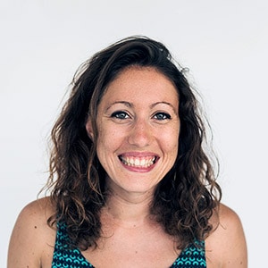 A woman with wavy brown hair and a green patterned sleeveless top smiles at the camera against a plain white background.