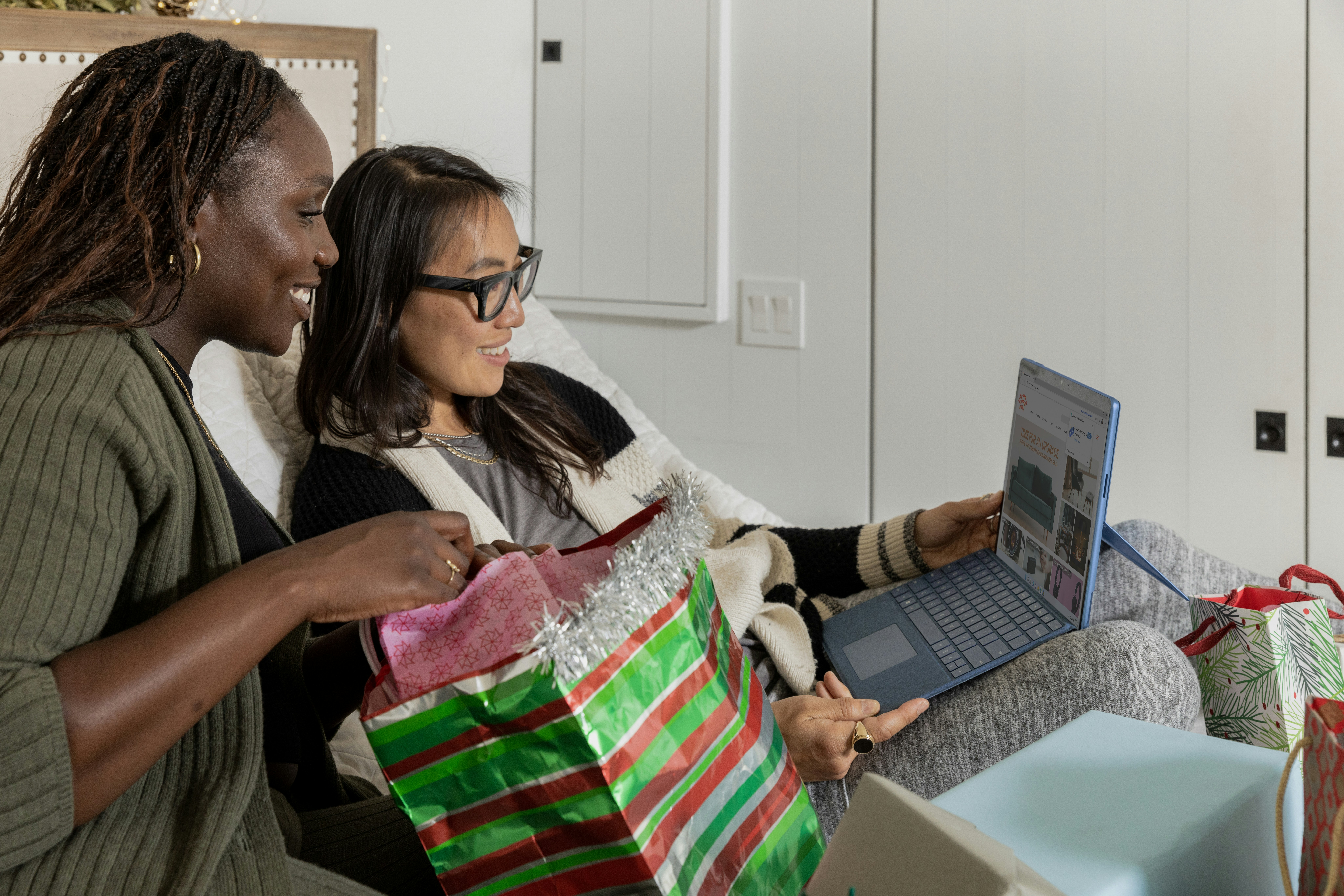 Two women sit on a couch looking at a laptop, with one holding a holiday gift bag and the other a wrapped present, discussing ESP for Small Business strategies. They are surrounded by festive decor, creating a cozy atmosphere for planning their email list management.