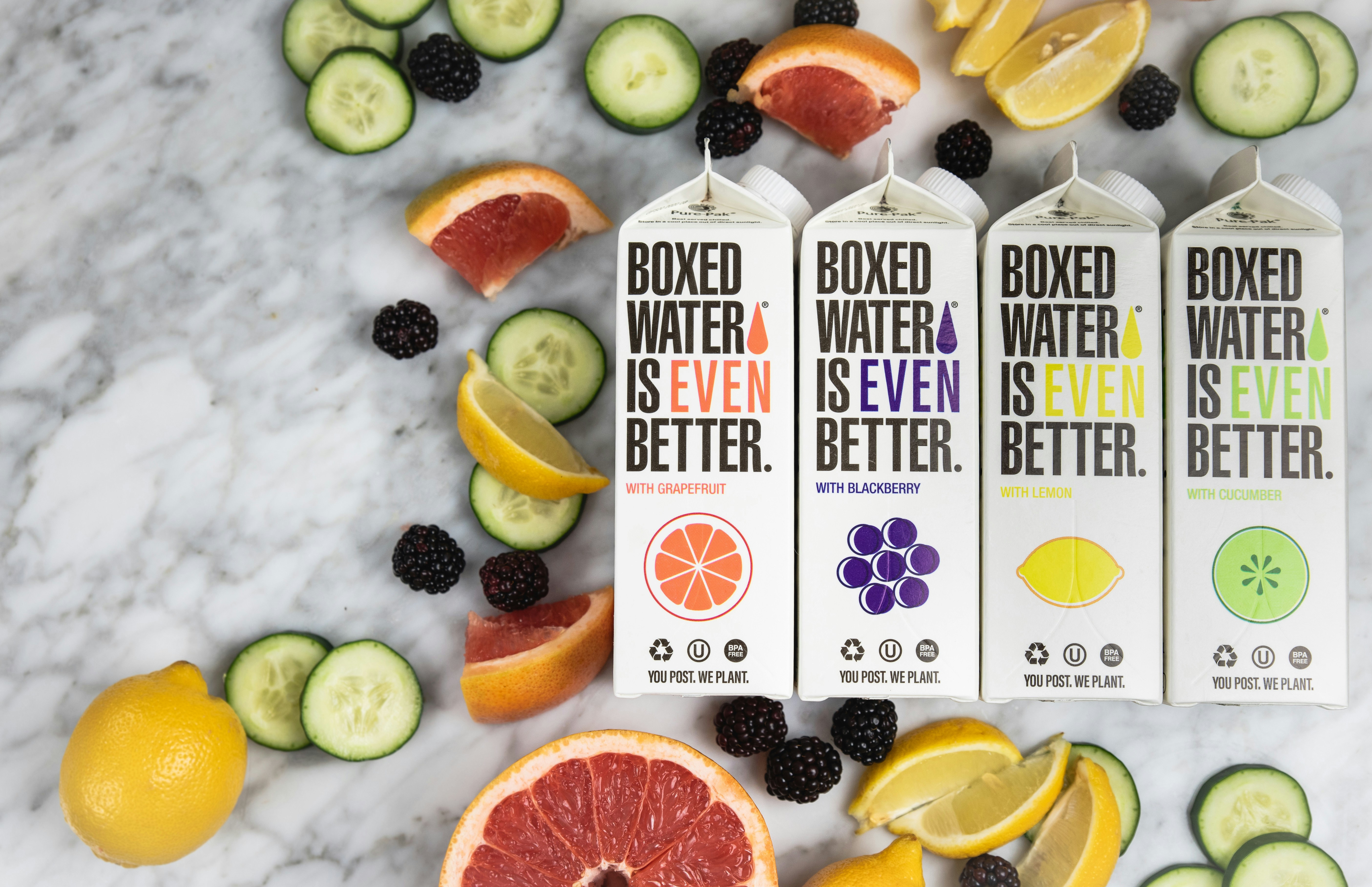 Four cartons of Boxed Water with different flavors, surrounded by assorted fresh fruit slices including grapefruit, lemon, cucumber, and blackberries, on a marble surface. Leverage email to share news about the refreshing options and engage effectively with your audience.