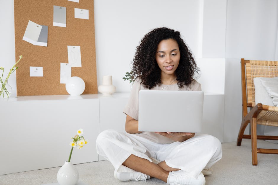 A person with curly hair, dressed in light casual clothing, sits on the floor in a room using a laptop. There is a corkboard with papers behind them and a small flower in a vase nearby. They seem focused on developing online marketing strategies for their next project.