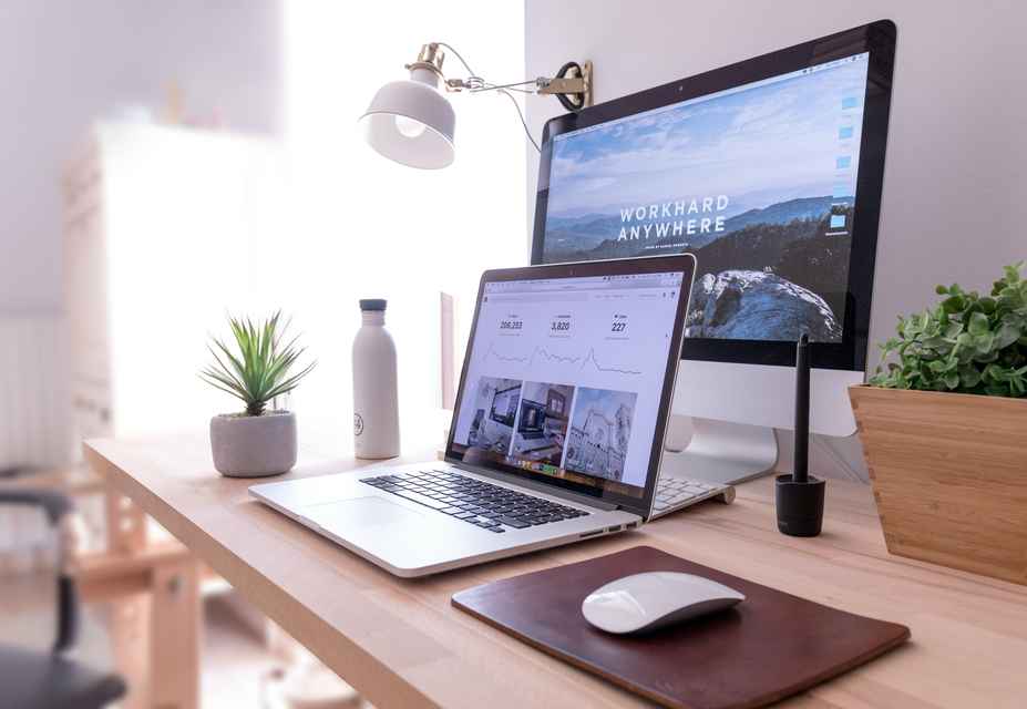 A work desk with a MacBook on a brown mousepad, an iMac displaying WORKHARD ANYWHERE, a potted plant, a water bottle, a graphics tablet, and a white desk lamp. The setup is perfect for focusing on User Experience (UX) design and responsive e-commerce integration.