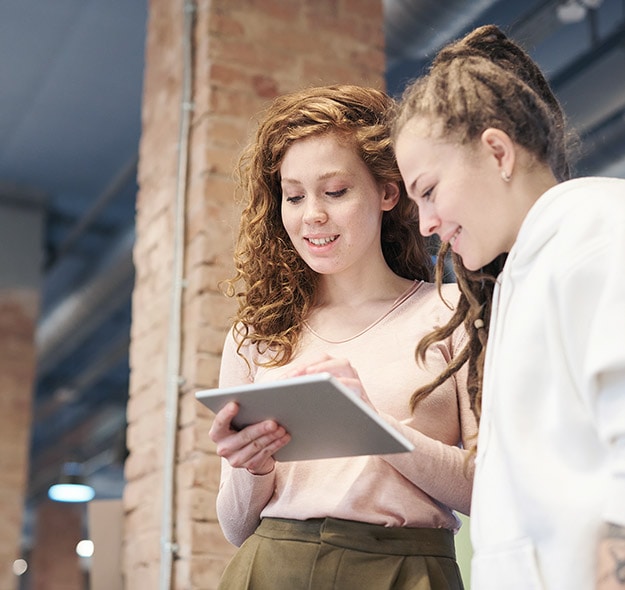 Two women standing indoors and looking at a tablet together. One has curly hair and is wearing a pink top, while the other has dreadlocks and a white hoodie.