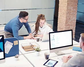 Three people in a modern office work at computers. One man stands while conversing with a seated woman about web design, while another person focuses on their desk's email service provider software. The office exudes a sleek, contemporary vibe.