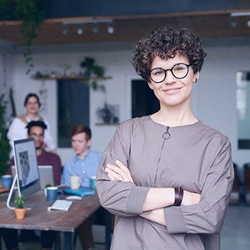 A person with short curly hair and glasses stands confidently with arms crossed in an office. Three people are working at a table behind them with computers and plants nearby, ready to handle any business inquiries.