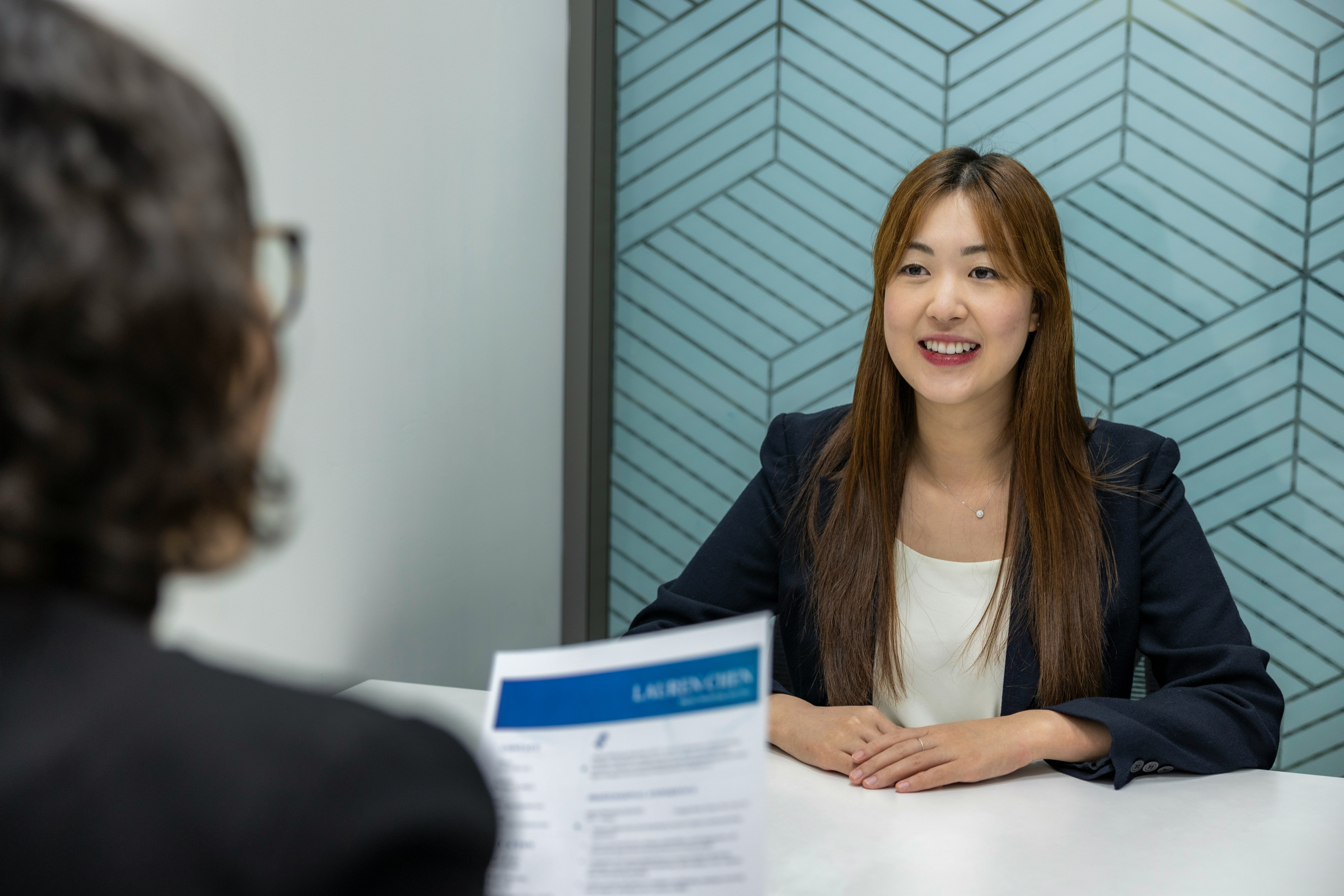 A woman with long brown hair sits at a desk, facing another person holding a resume during an interview in a modern office setting, discussing employer branding techniques.