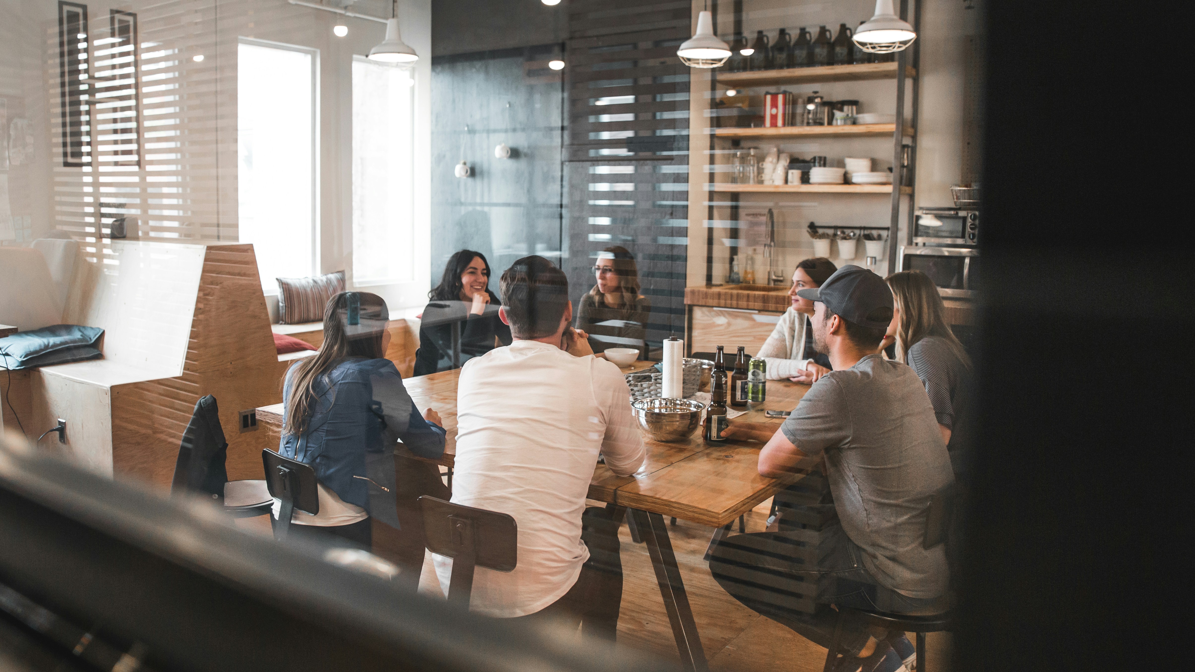 A group of people sit around a large wooden table in a modern kitchen, engaged in conversation about social media strategy. The room features shelves with various items and pendant lighting above the table.