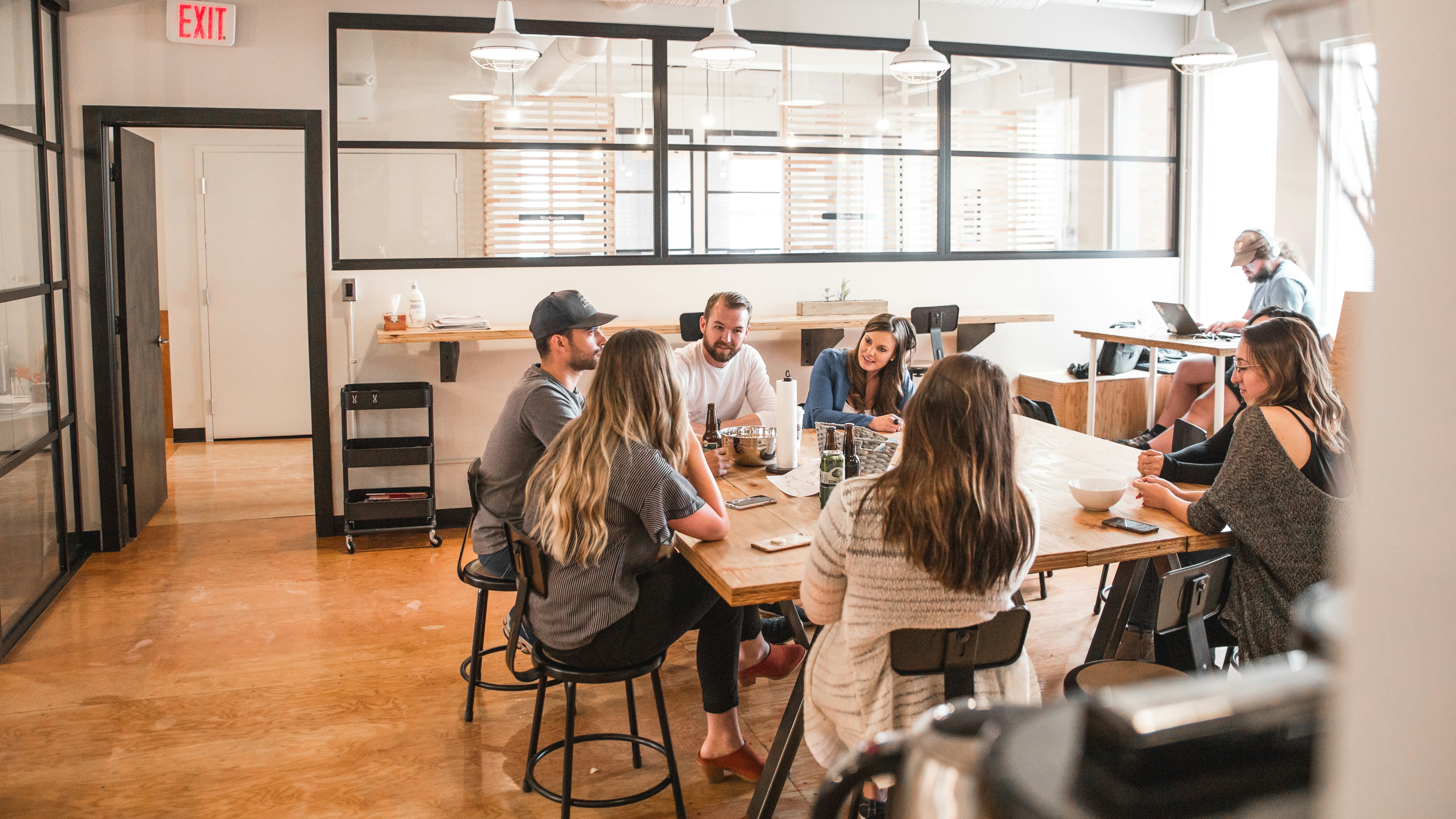 A group of people sit around a table in a well-lit office space having a meeting about content creation, while one person works on a laptop in the background.