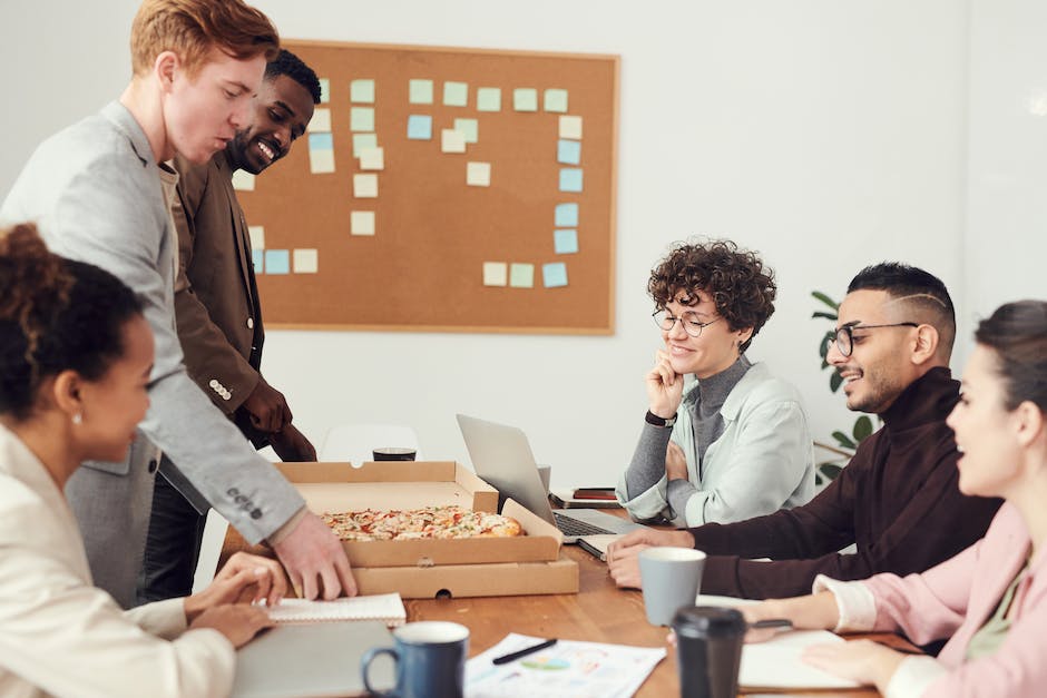 A group of five people are gathered around a table with a laptop and an open pizza box. They are brainstorming affordable web design solutions in a meeting room, featuring a corkboard covered in sticky notes in the background.